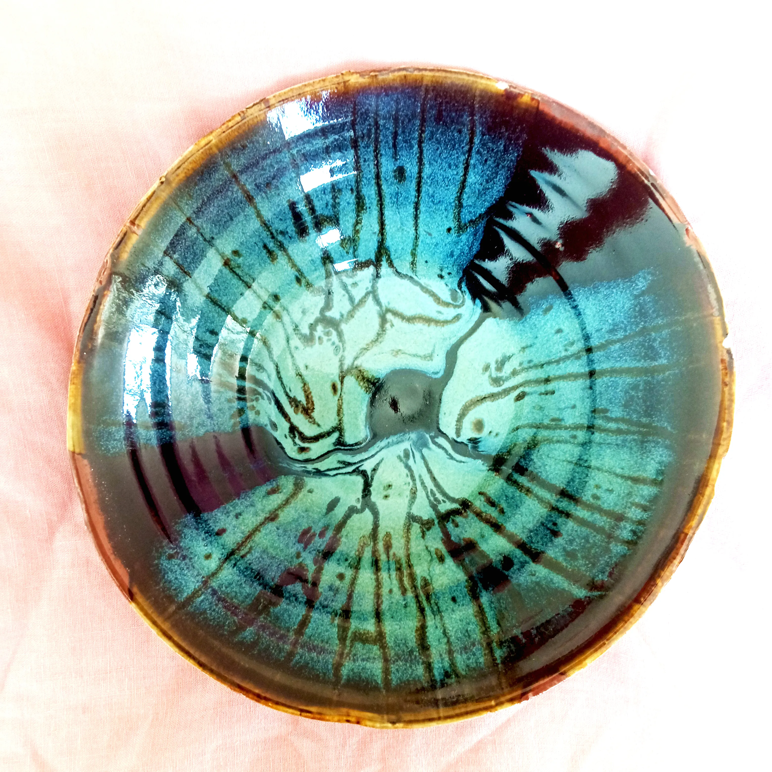 Round hand-built ceramic bowl glazed in turquoise and teal with dark radiating drip lines and an amber rim, seen from above on a pink cloth.