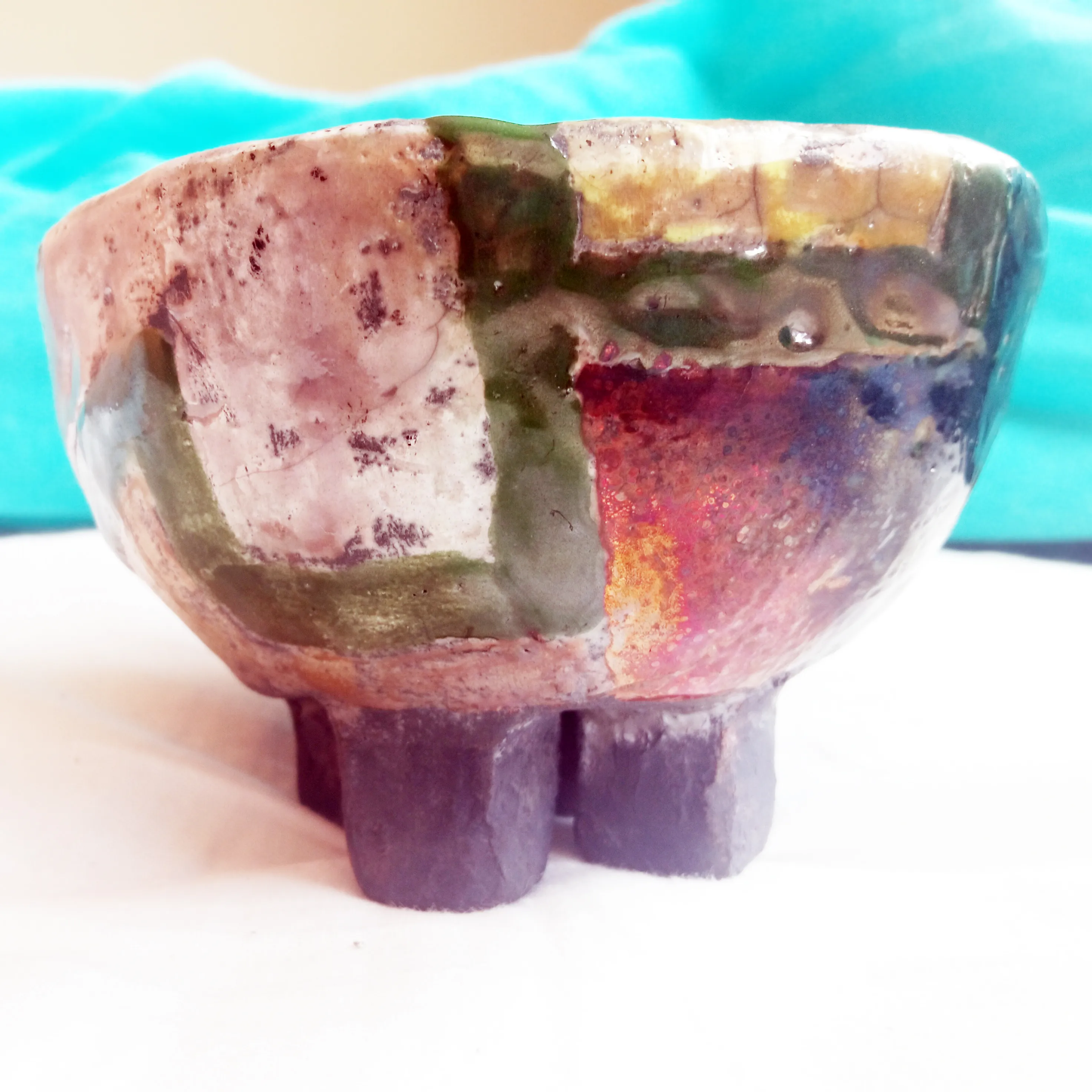 Footed raku bowl standing on short block legs, its surface a patchwork of pink, green, red and iridescent copper glaze.
