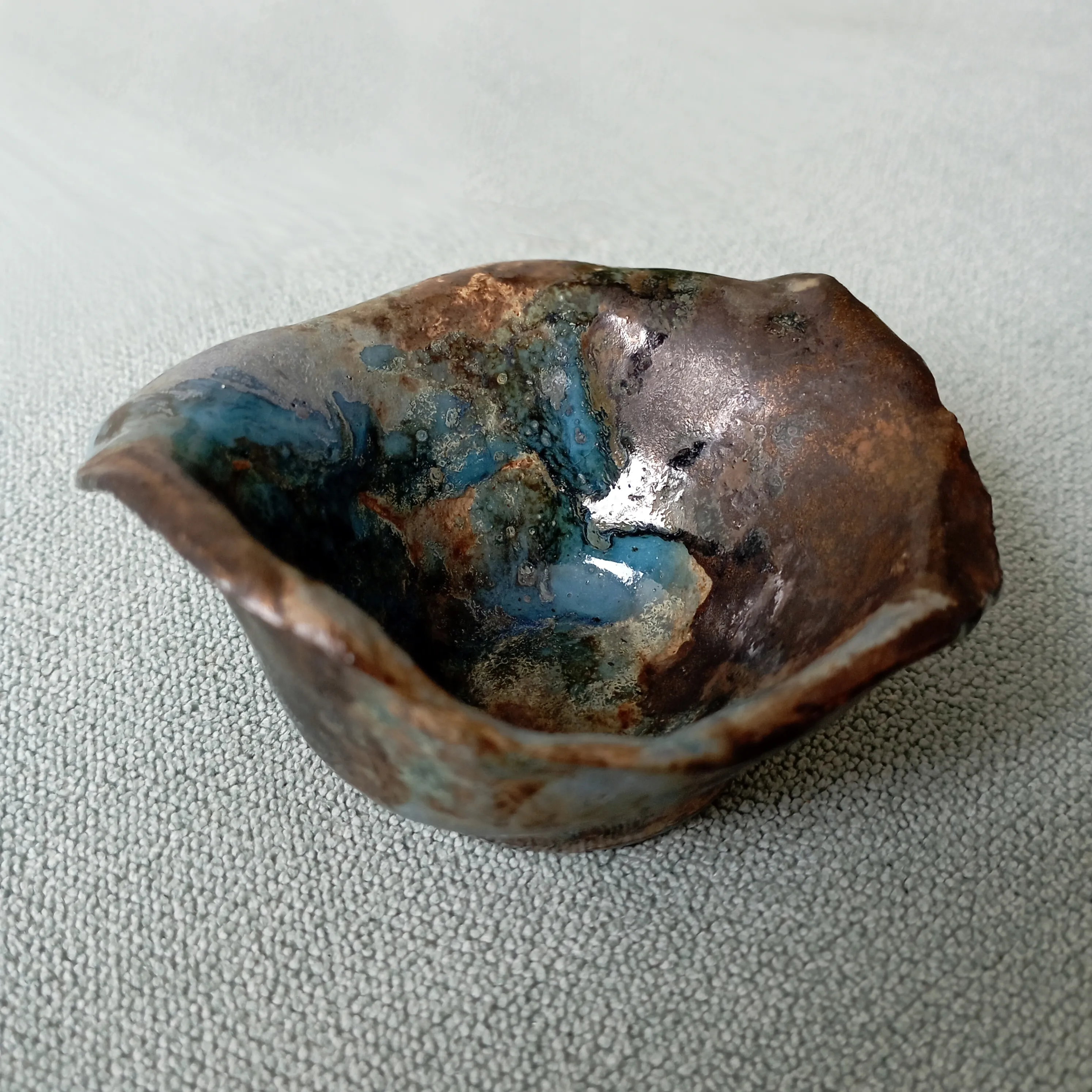 Small pinched raku bowl in metallic copper and bronze tones with a turquoise-blue glazed centre, resting on a grey surface.
