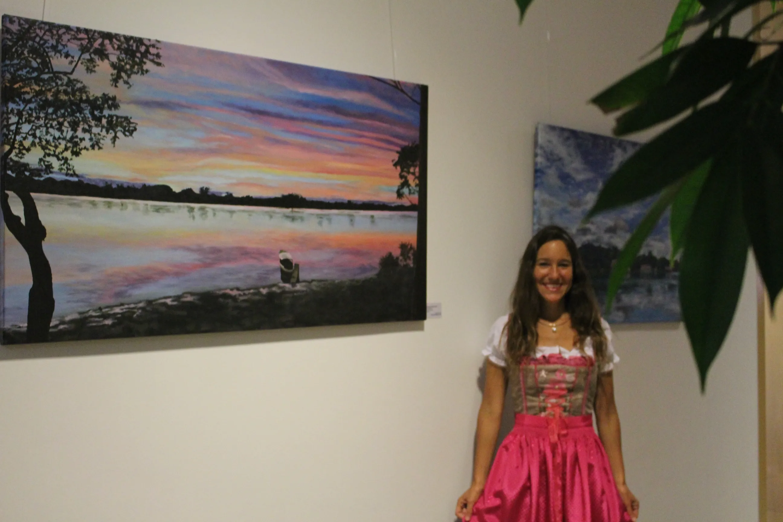 Ximena Heraud in a pink dress standing beside her large sunset lake landscape painting hung in the Goethe Gallery, Frankfurt