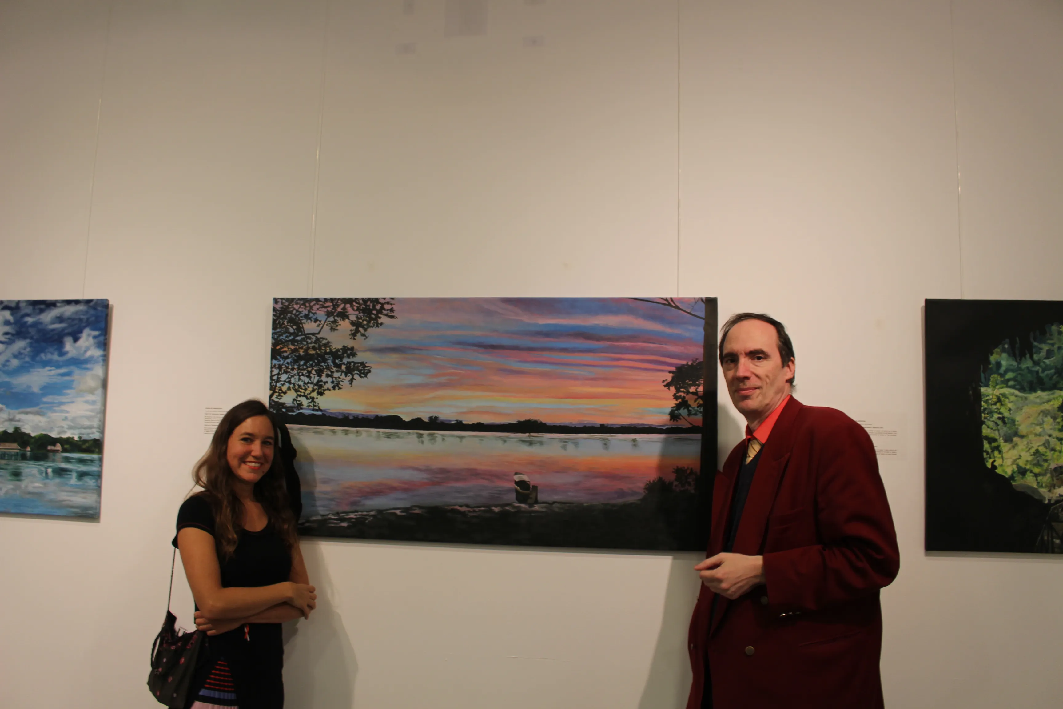 Ximena Heraud standing with a host beside her panoramic sunset landscape painting at the Bucharest exhibition, other works visible