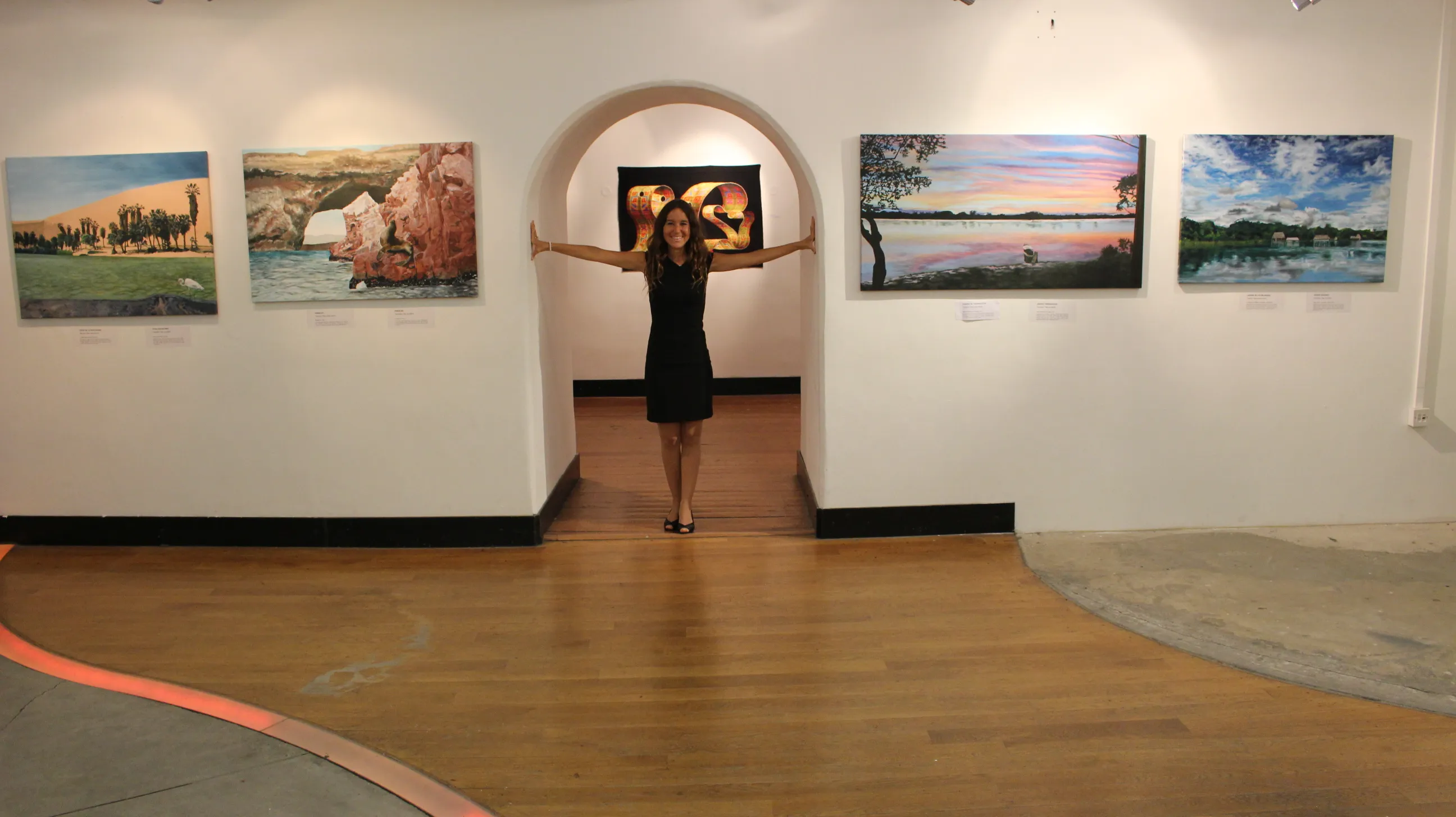 Ximena Heraud with arms outstretched framed by a stone archway in the Prague gallery, her landscape paintings lining both walls