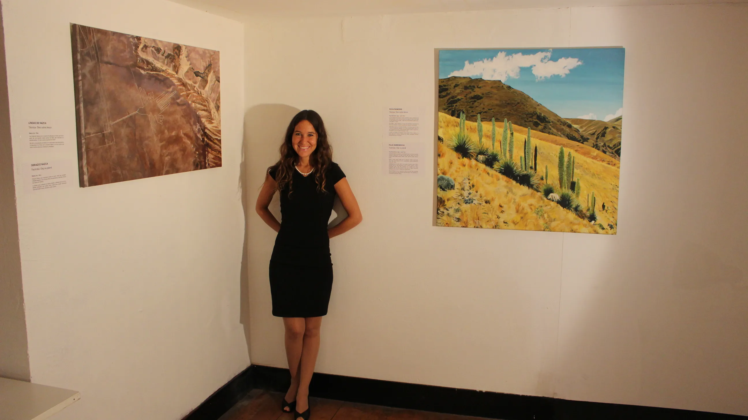 Ximena Heraud in a black dress beside two of her paintings, an abstract and a cactus landscape, on the wall in Prague