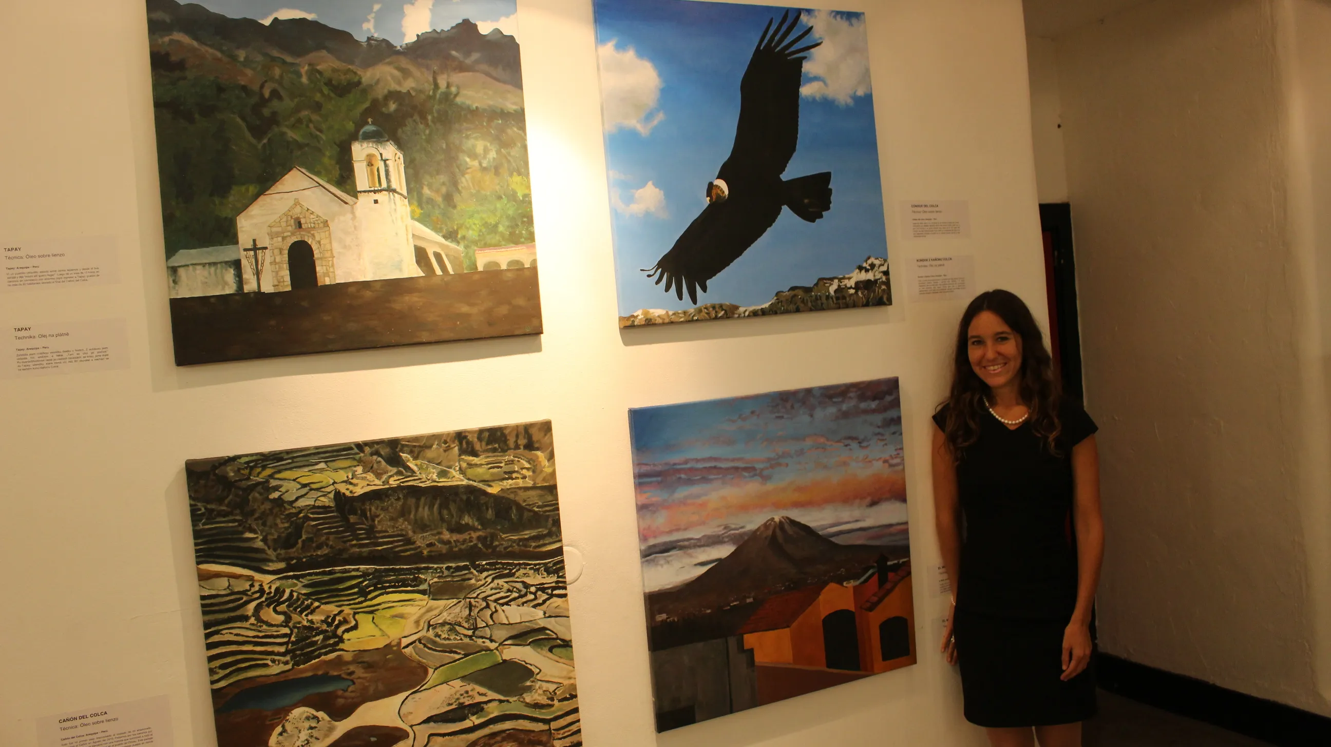 Ximena Heraud in a black dress beside her Recorriendo Peru landscape series, a white chapel, a condor and a volcano, hung in the Prague gallery