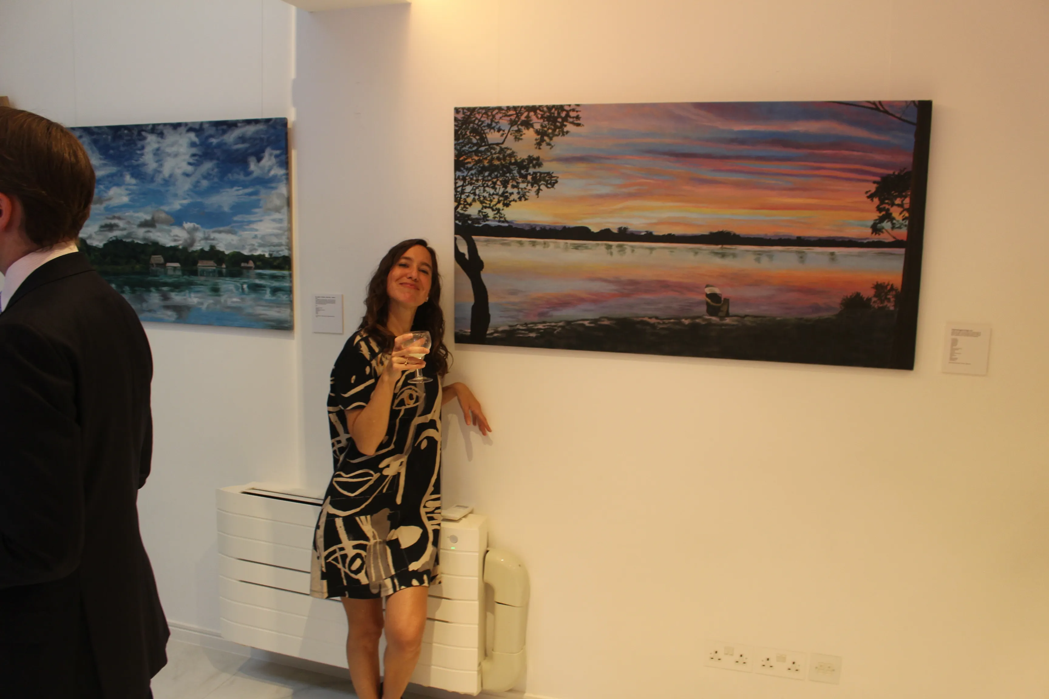 Ximena Heraud standing beside her sunset lake landscape painting on the wall at the Embassy of Peru in London
