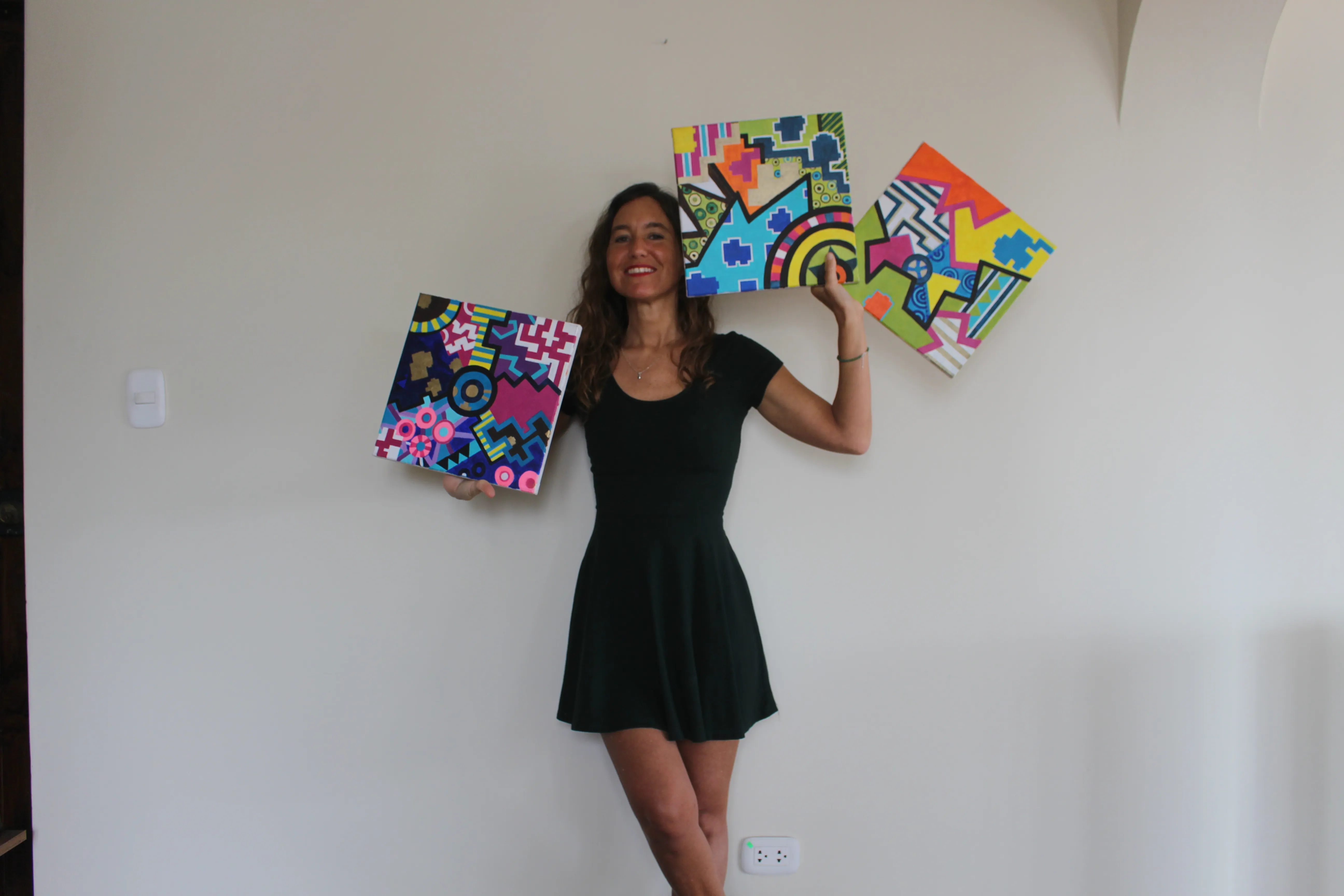 Ximena Heraud in a black dress holding two of her vivid geometric paintings while more hang on the wall at the Conexiones show in Lima