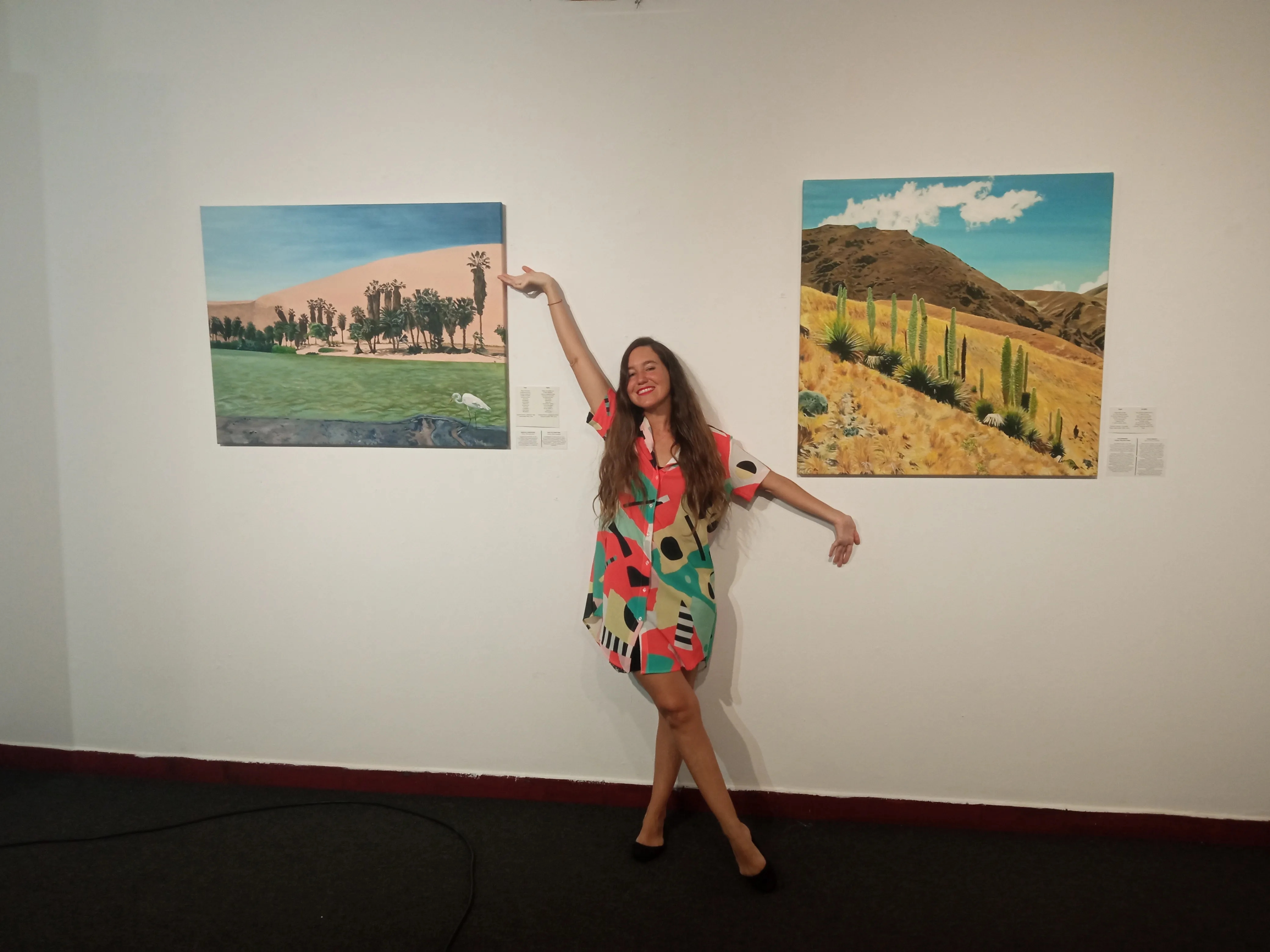 Ximena Heraud in a bright patterned dress with arm raised beside two landscape paintings at the Athens exhibition