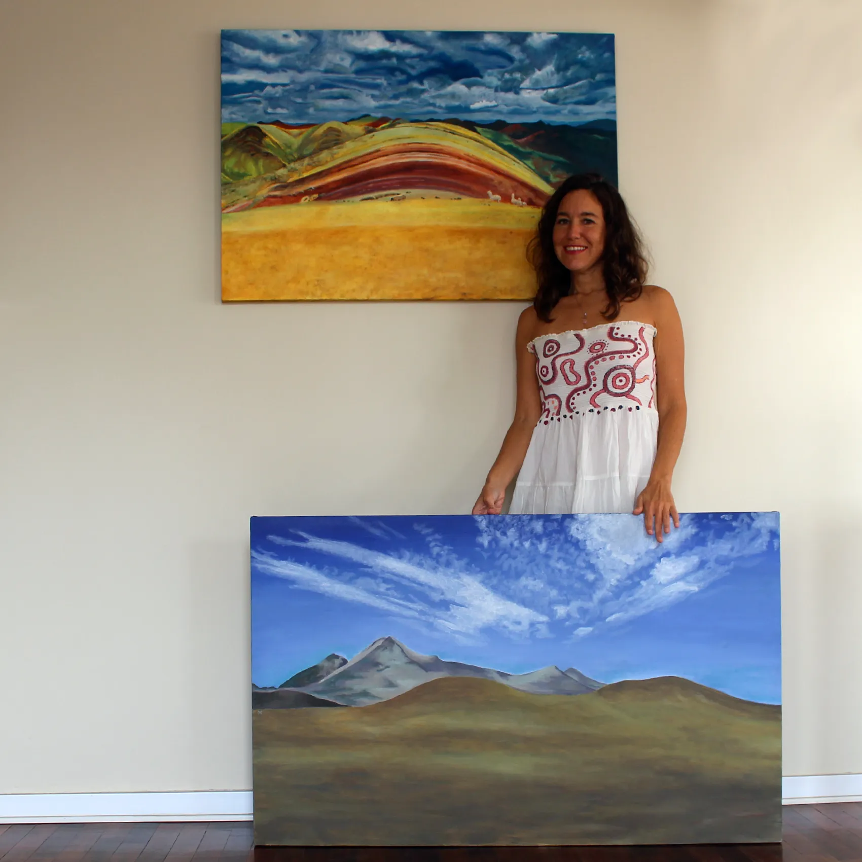 Ximena Heraud standing between two of her Peruvian landscape paintings, one hung and one resting on the floor, in a bright studio space