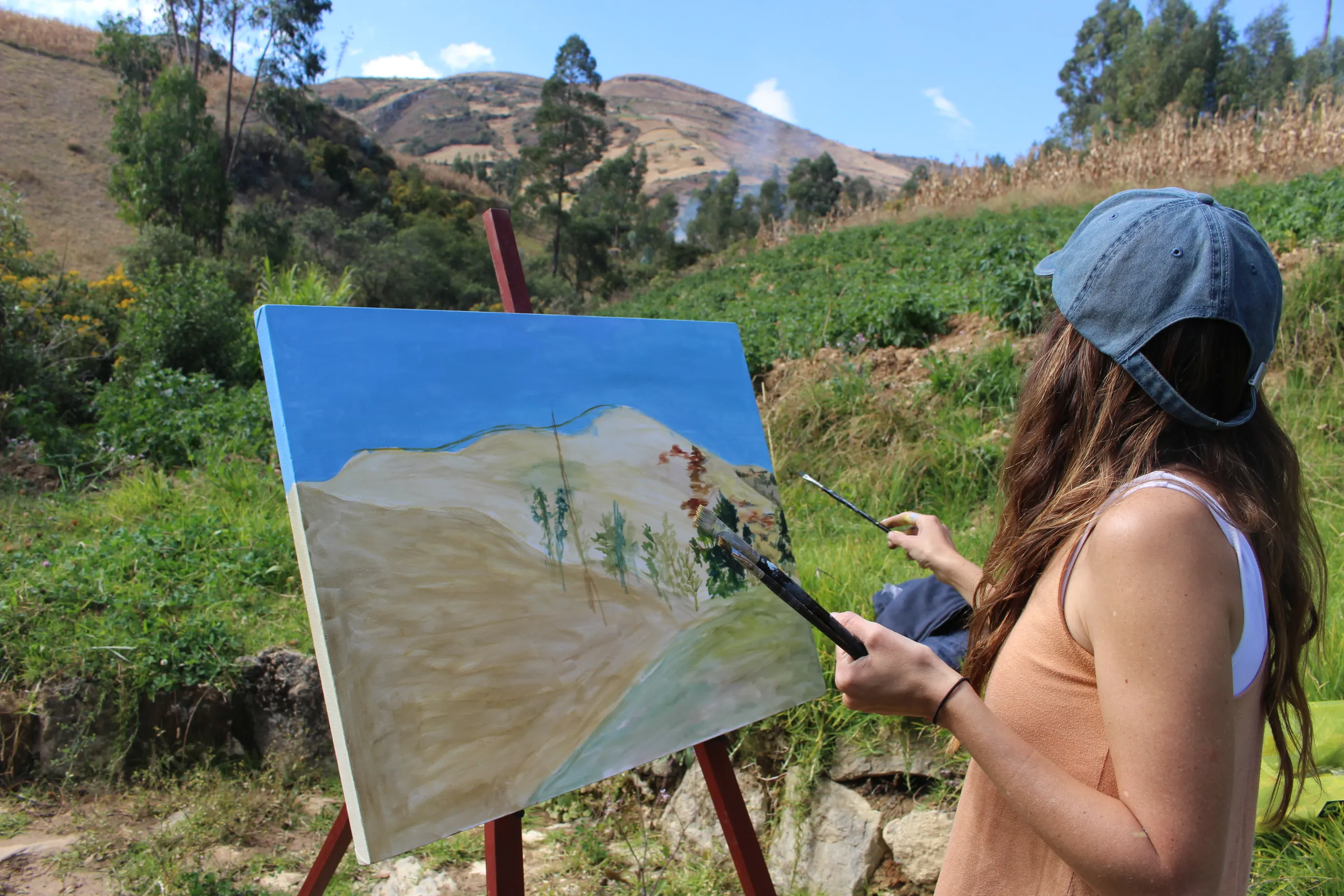 Ximena Heraud in profile wearing a cap, brush in hand, painting a landscape on canvas at an easel in the green Peruvian highlands of Cajamarca
