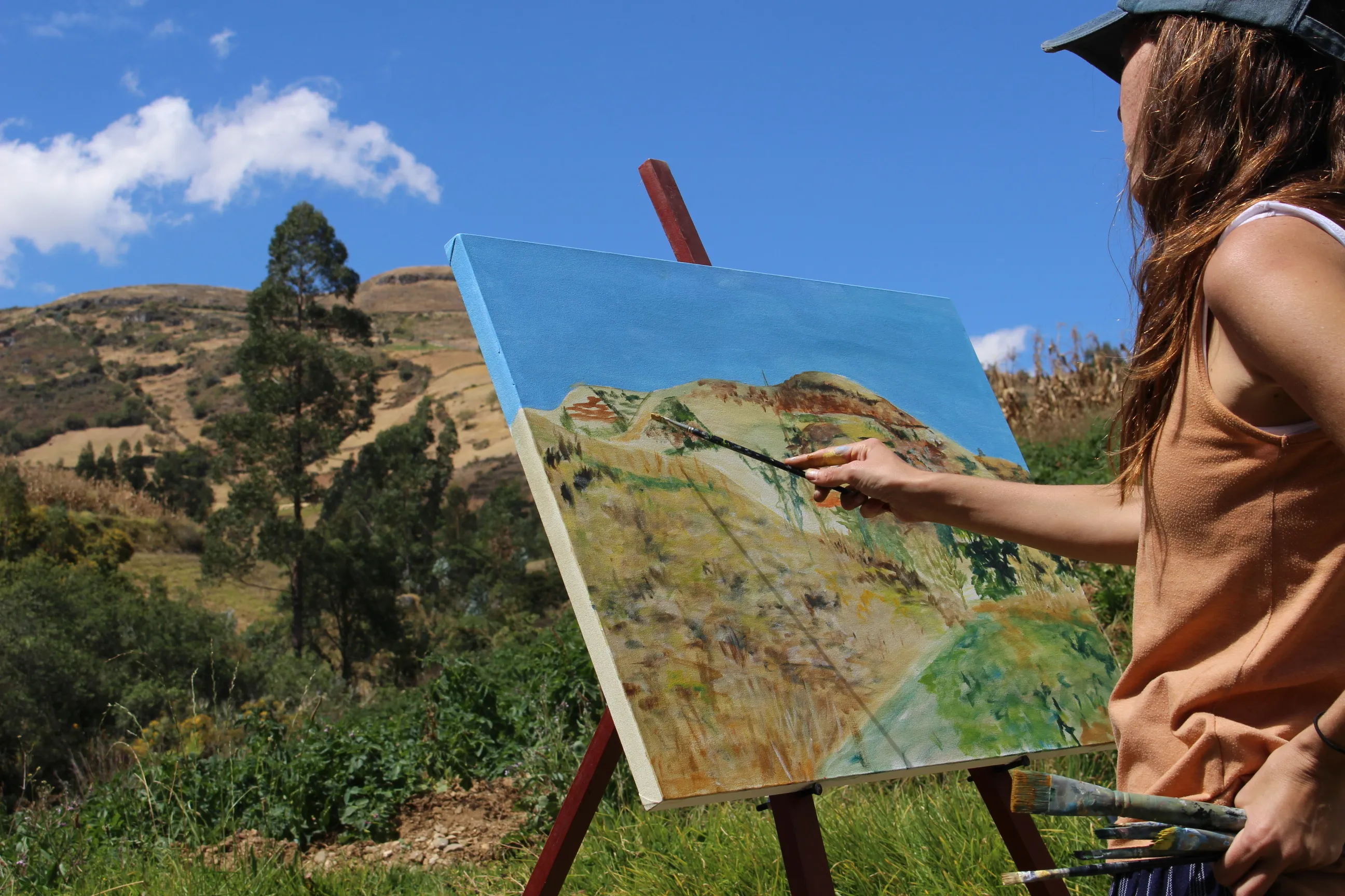 Ximena Heraud applying paint to a canvas whose sky and hills mirror the actual Cajamarca valley behind her easel