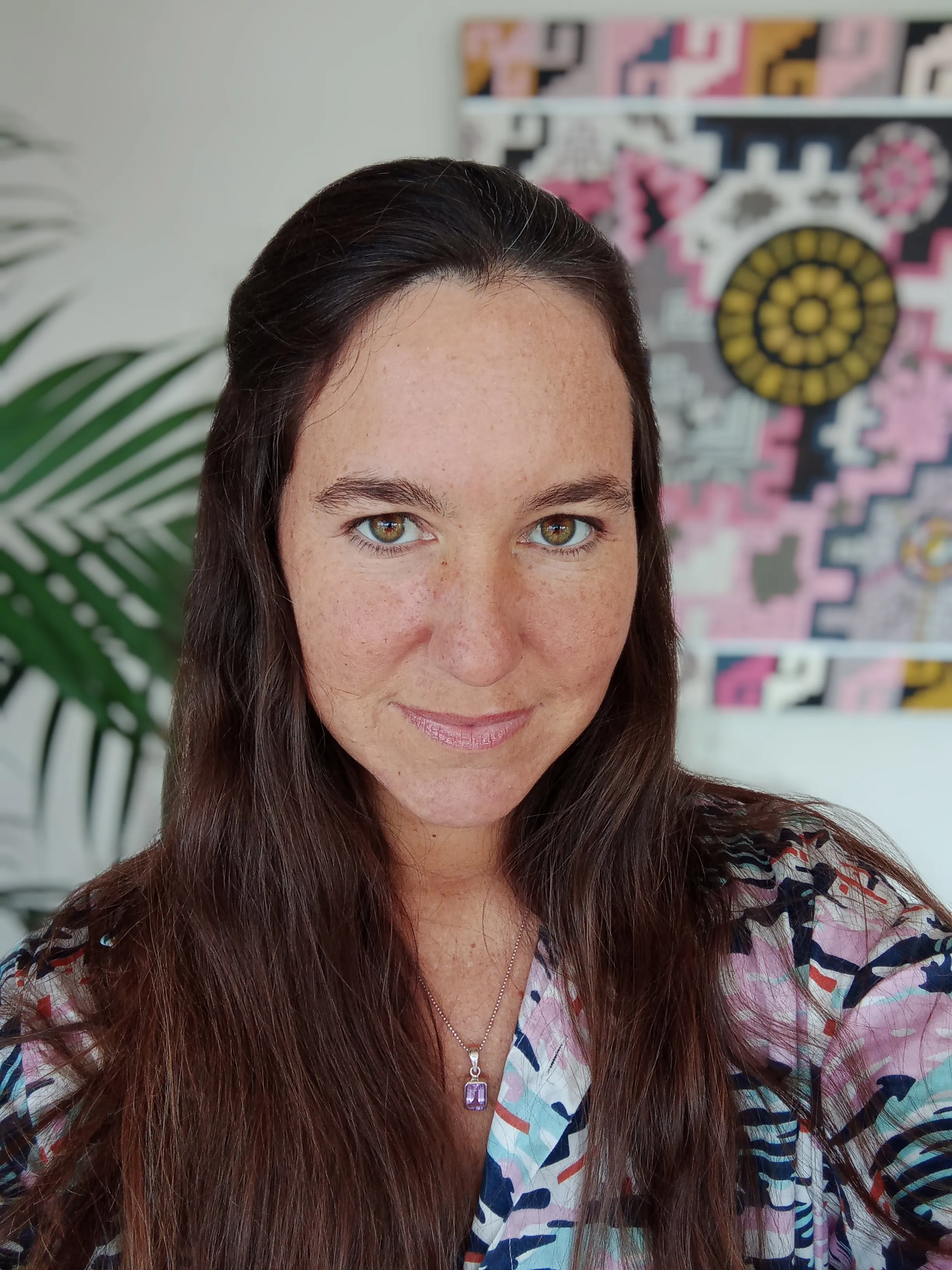 Close, well-lit portrait of artist Ximena Heraud, long dark hair, looking directly at the camera in front of one of her colorful geometric works