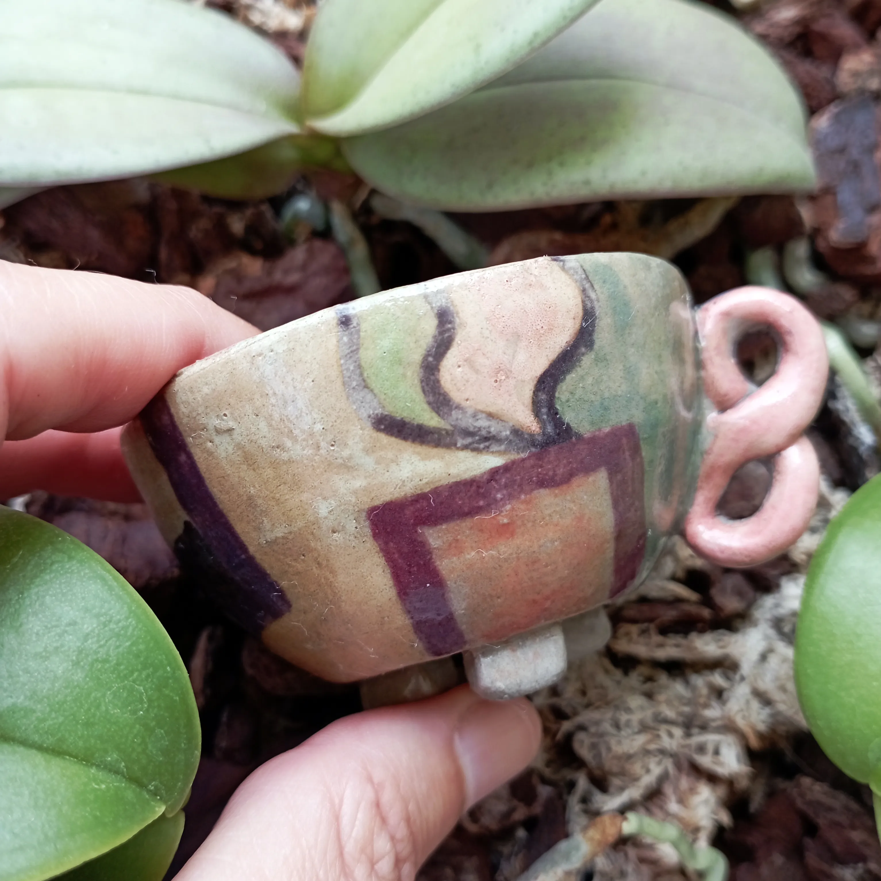 Small footed cup in cream clay painted with maroon, green and purple geometric shapes, with a looping pink pretzel-shaped handle.