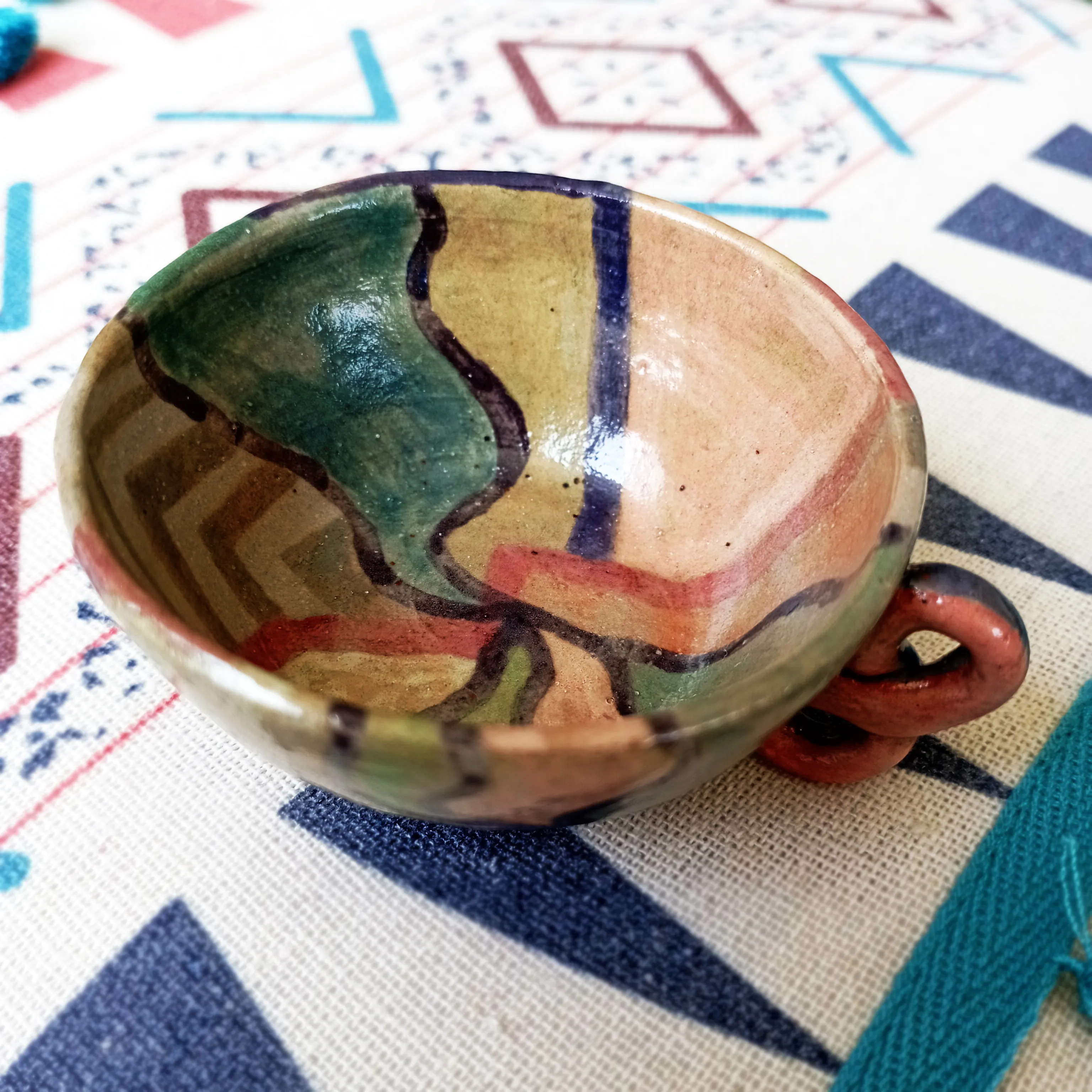 Glazed ceramic cup seen from above showing an interior painted with green, pink, red and blue geometric fields and a small handle, on a patterned Andean textile.