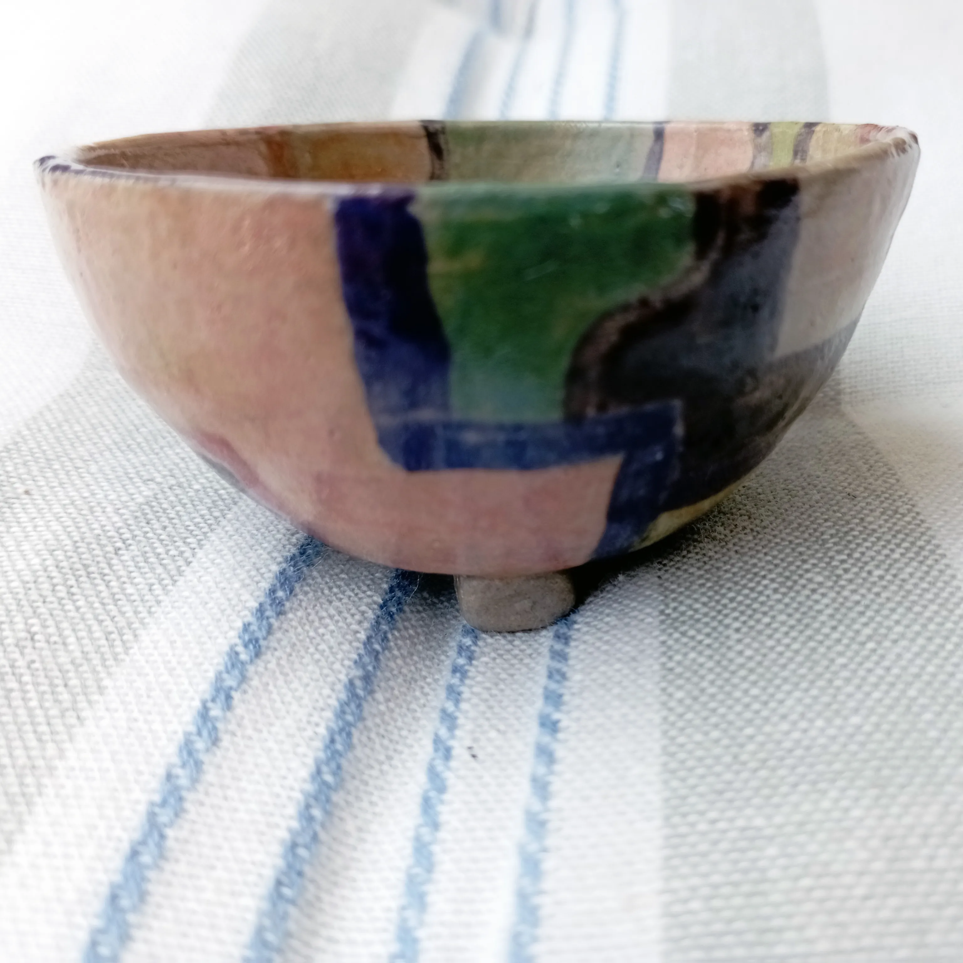 Footed ceramic cup viewed from the side, painted in pink, cobalt blue and green blocks, resting on a blue-striped cloth.