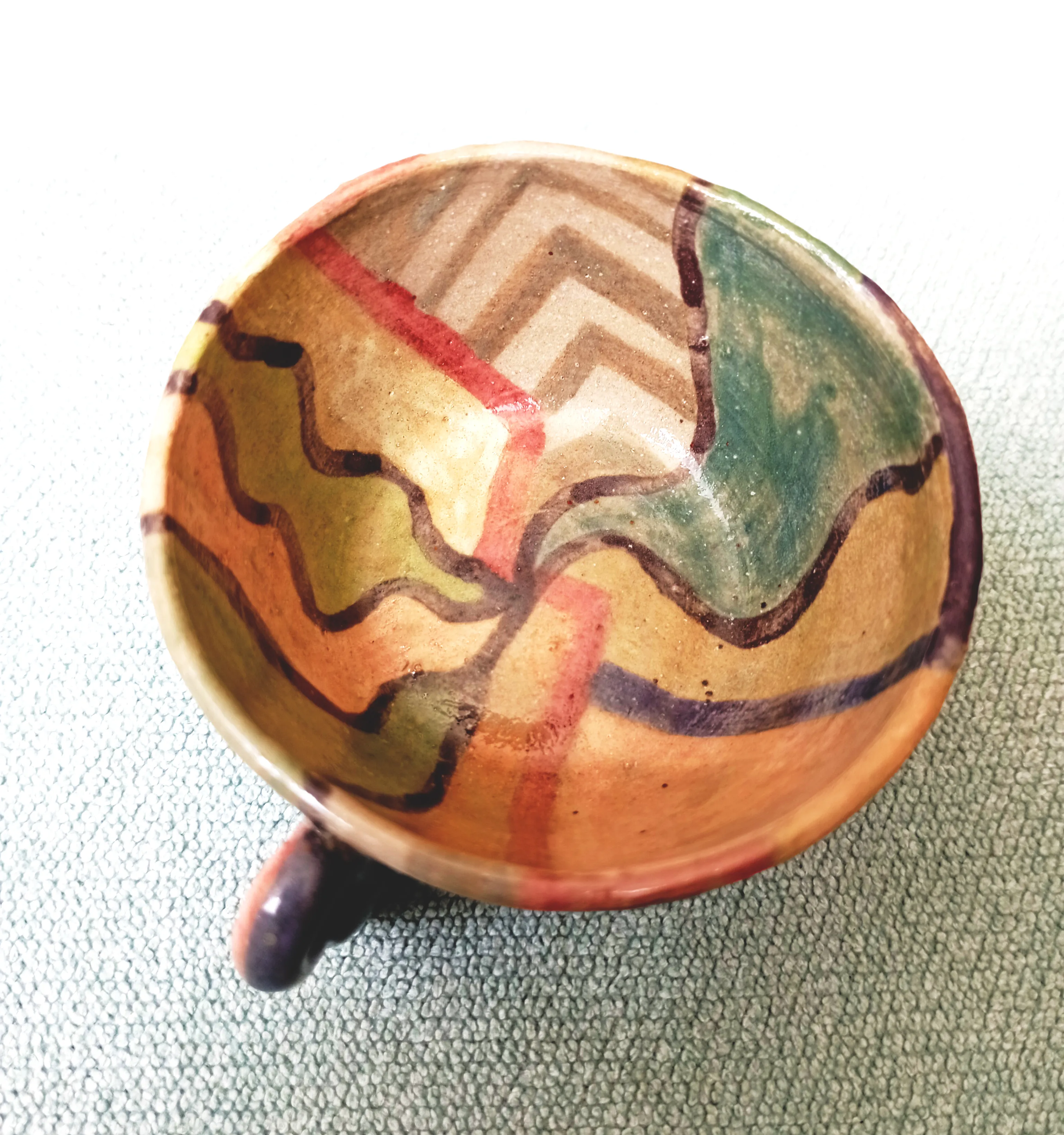 Earth-toned ceramic cup seen from above, its interior painted with wavy brown lines, red bands and a green wedge over a chevron pattern.