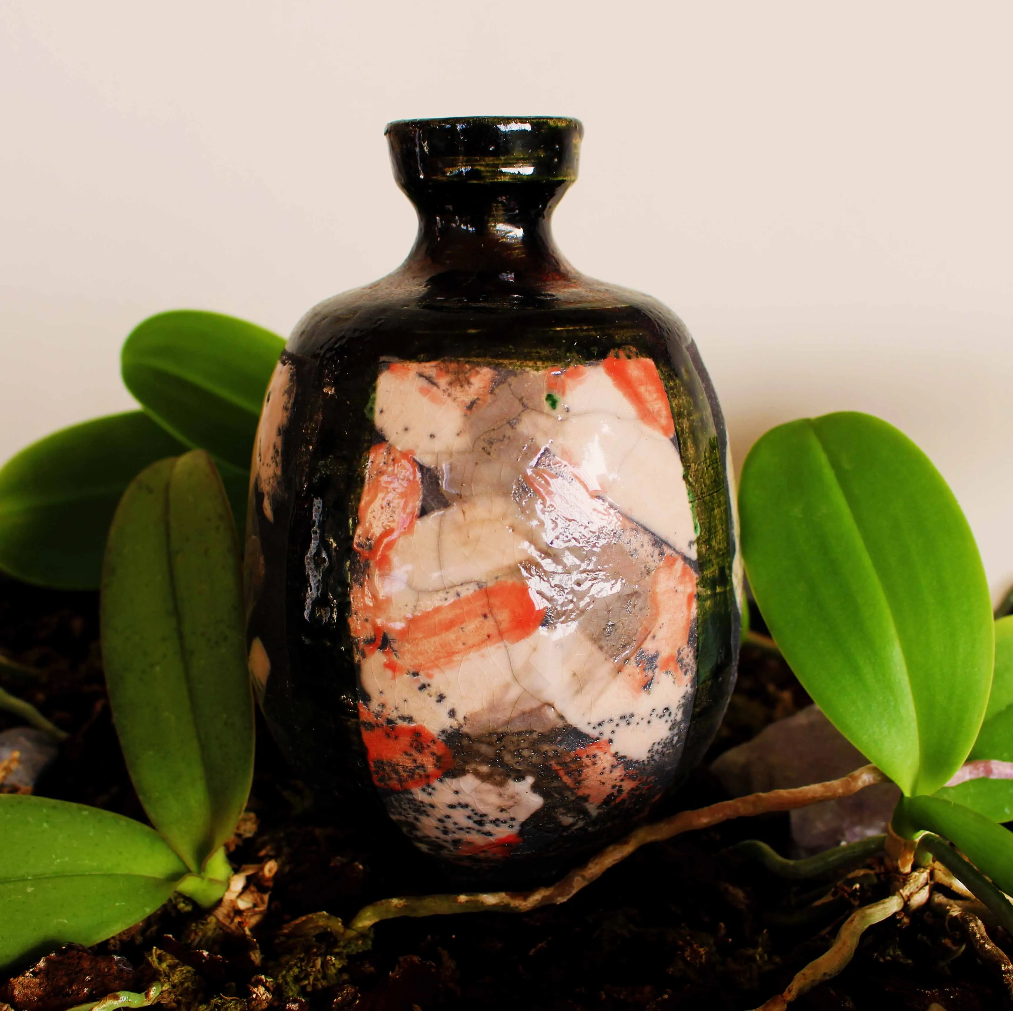 Small raku bottle vase with a dark green-black glaze and crackled panels of cream and orange, standing among orchid leaves.