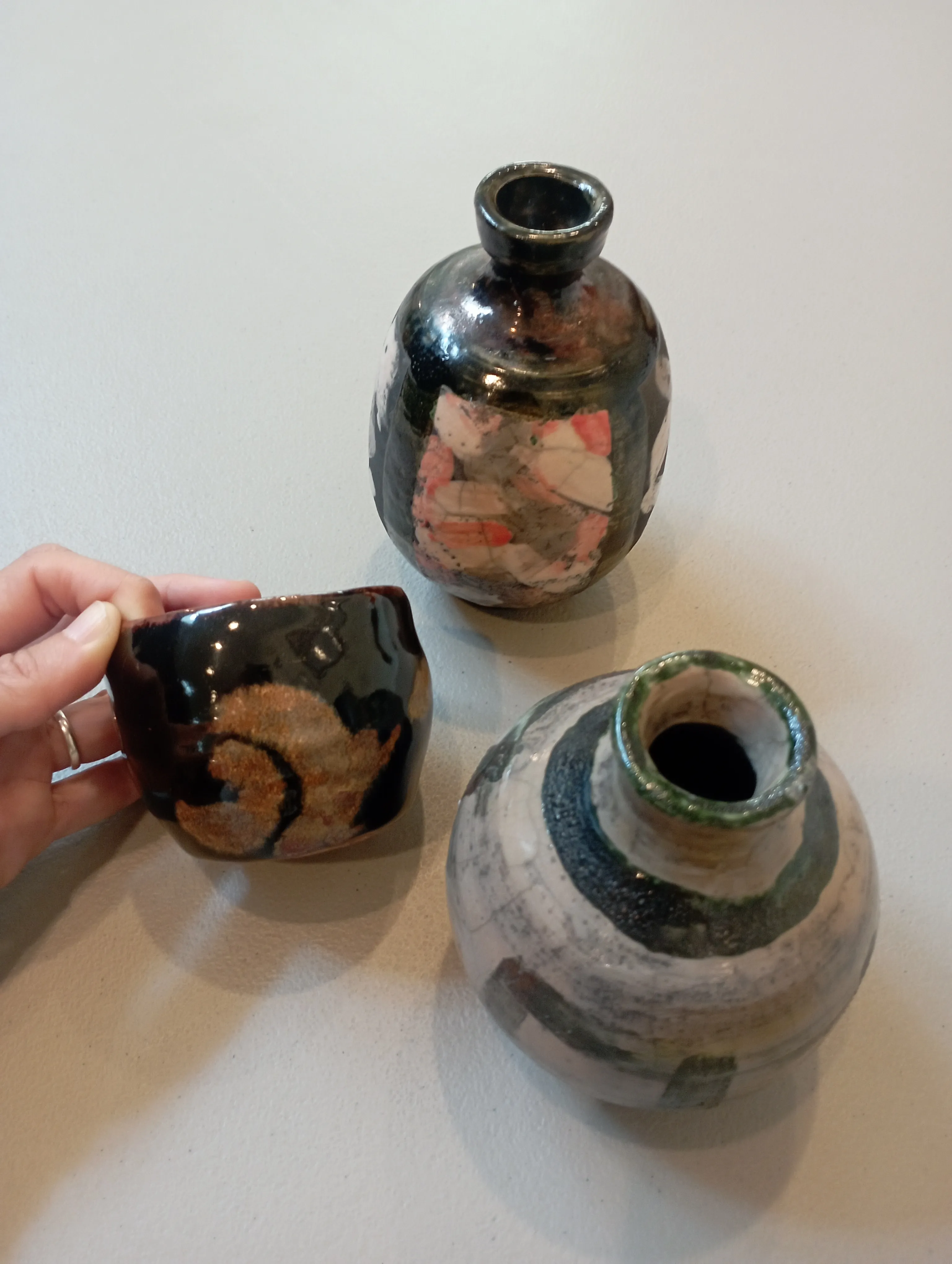 Three raku bottle vases photographed from above against a pale wall, one dark with orange patches, one white with green crackle, and a smaller dark vessel held in a hand.